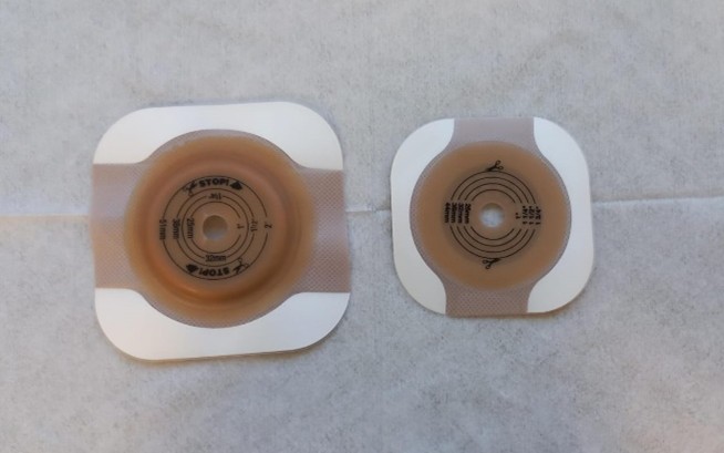 Bases adesivas para bolsas de colostomia em dois tamanhos diferentes. Dois modelos de base adesiva para bolsa de colostomia, posicionados lado a lado sobre uma superfície branca. A base da esquerda é maior que a da direita, ambas com marcações circulares para recorte ao redor de um orifício central.